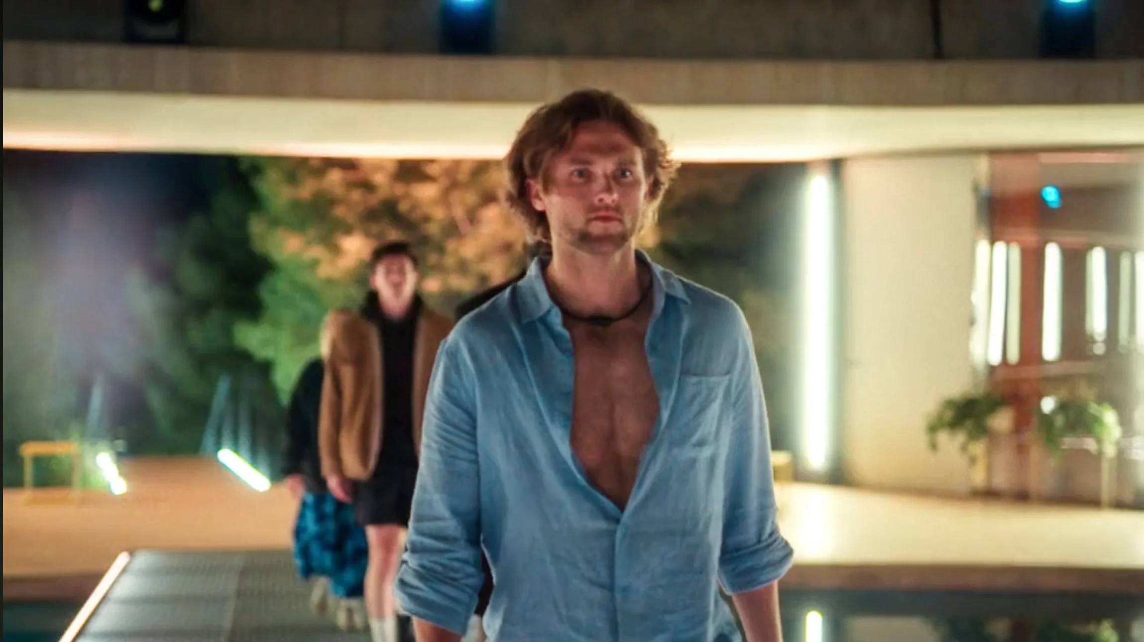 A man with long, curly hair and a beard stands outdoors at night with a serious expression, wearing a partially unbuttoned blue shirt. In the background, another person with dark hair and a brown jacket is walking behind him, with blurred greenery an