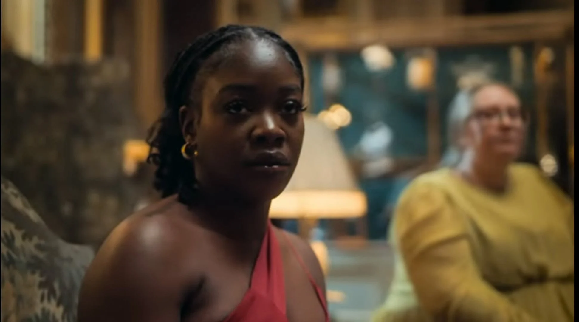 A woman with dark skin and braided hair, wearing a red dress and gold hoop earrings, sitting in a warmly lit room with blurred background.