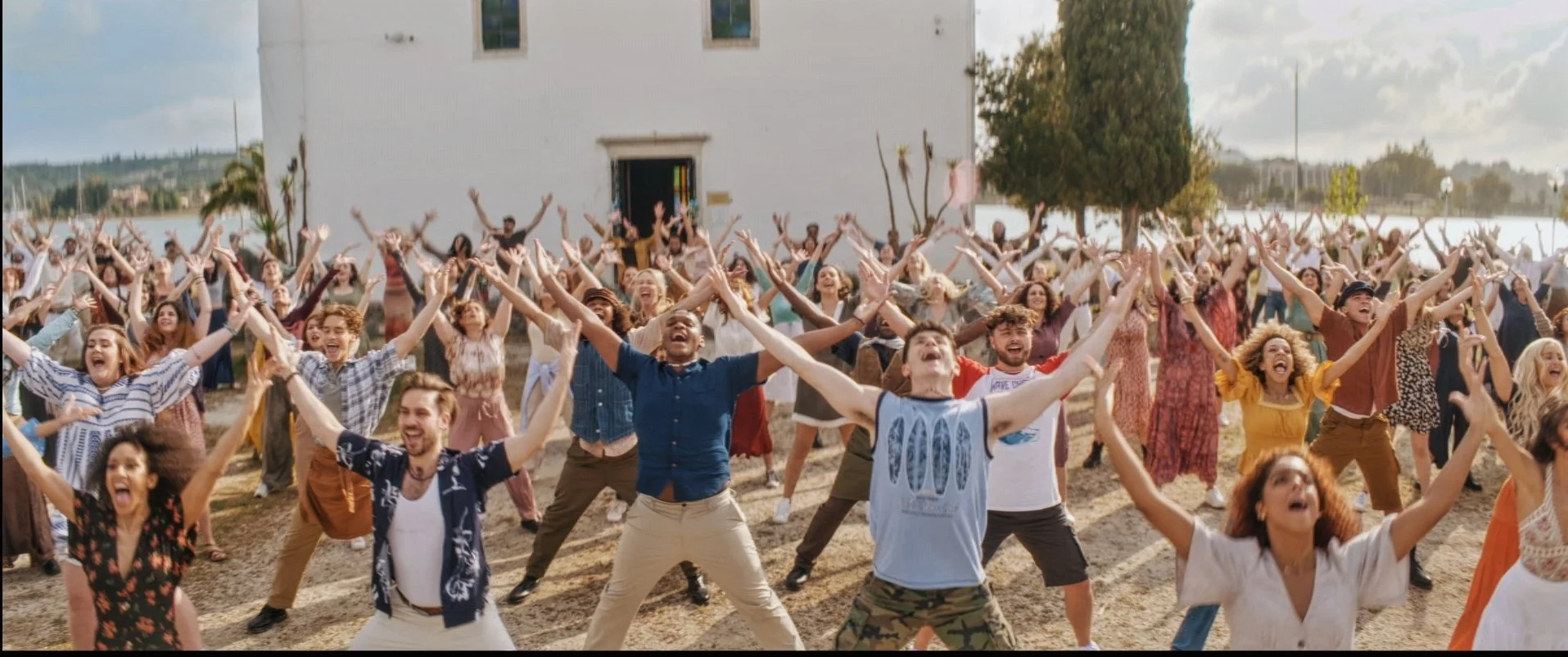 A large group of diverse people outdoors, smiling and raising their hands in celebration in front of a white building and trees.