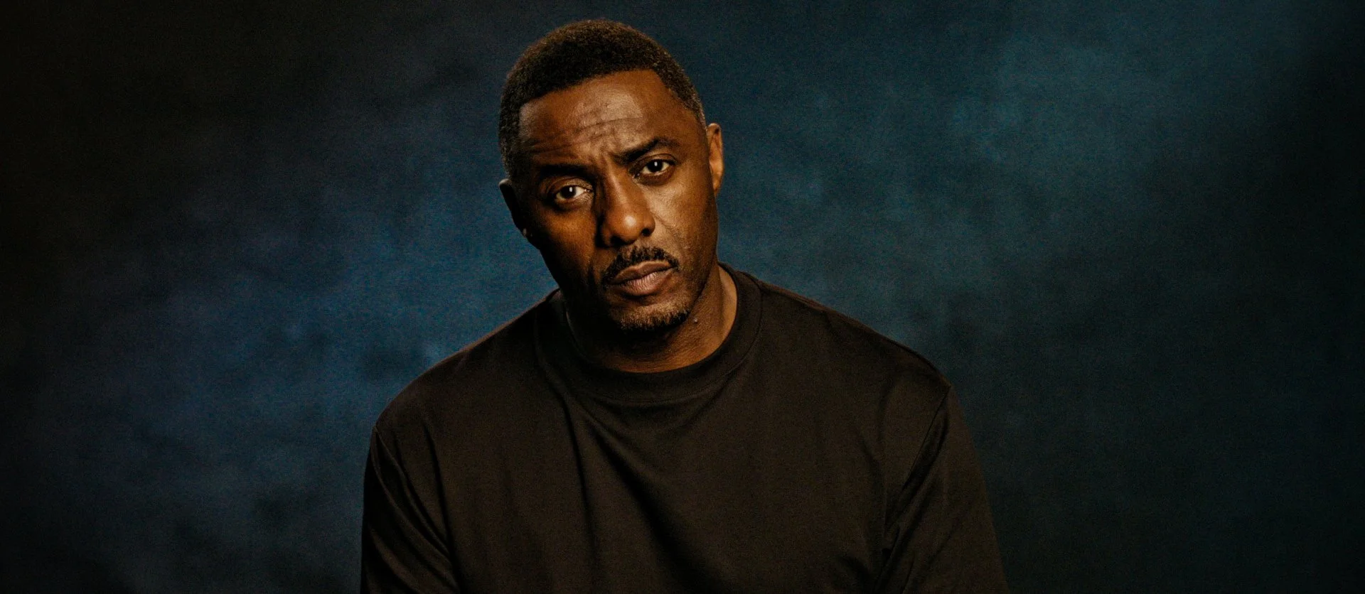 Portrait of a man with a serious expression, wearing a black shirt, against a dark background.