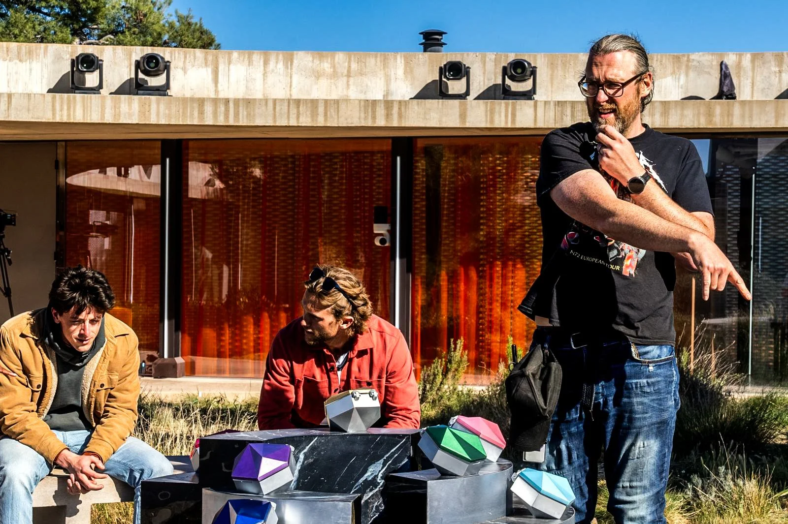 Three men outdoors near modern building with large glass windows, one standing and pointing, two sitting, near colorful geometric sculptures.