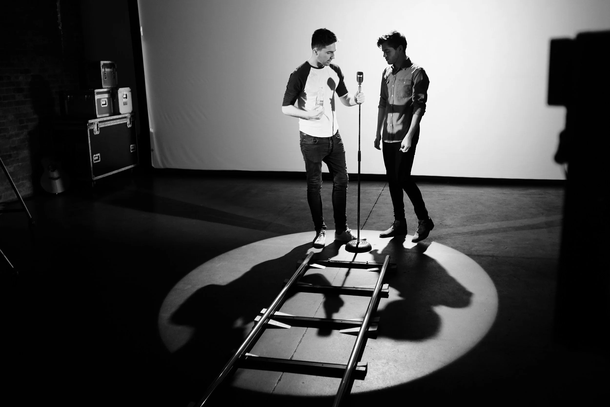 Two young men stand on stage, one holding a water bottle and the other looking at a microphone stand, with a spotlight on them and a ladder on the floor, in a black-and-white photo.