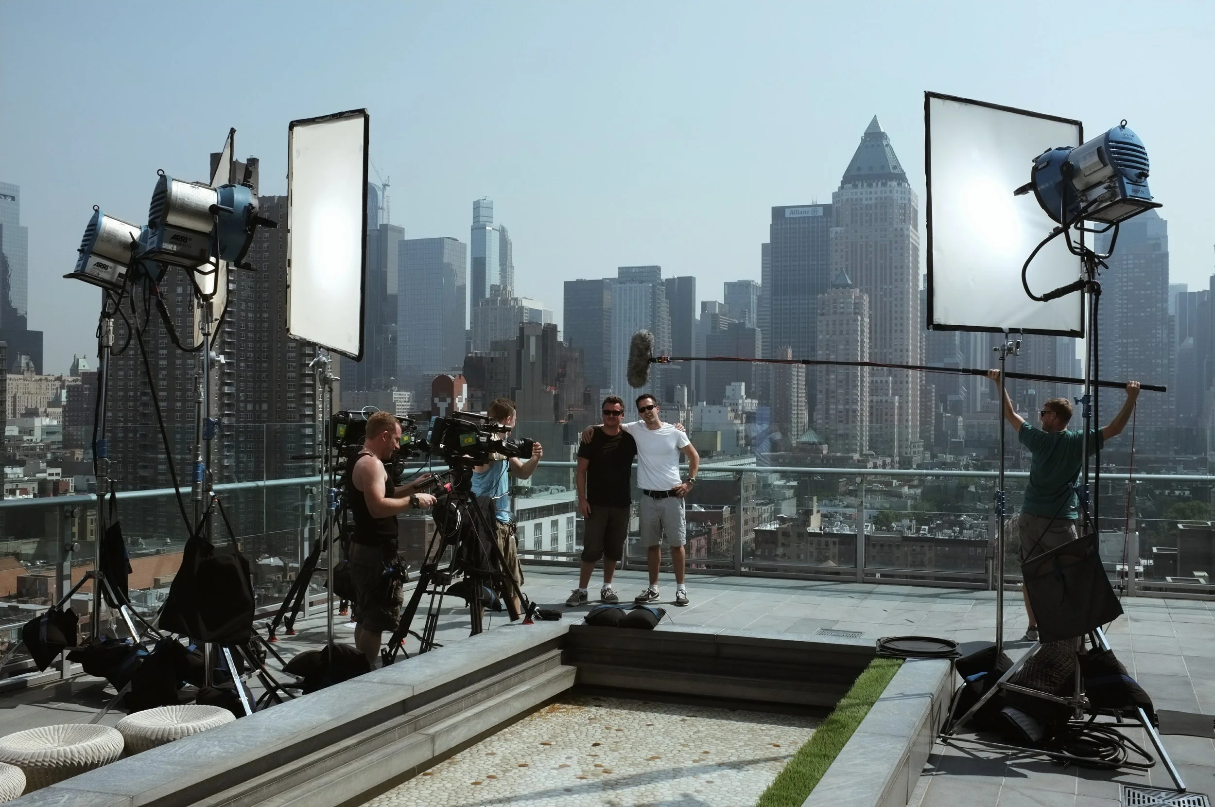 Filming crew on rooftop with city skyline in background, two men posing for camera, large lights and equipment around.