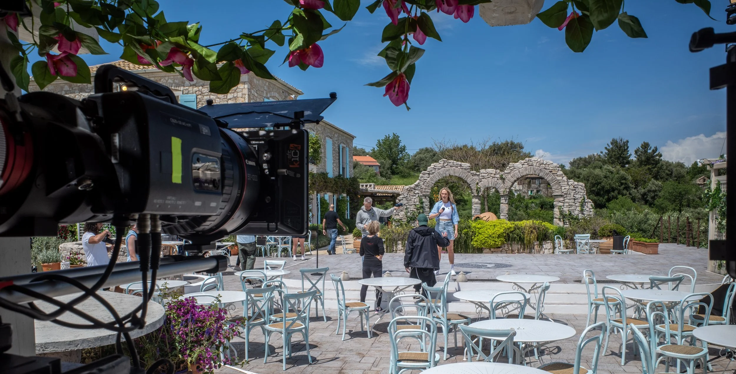 A film crew is filming outside on a patio with white tables and chairs, with a stone building and arches in the background, under a bright blue sky, with a camera in the foreground.