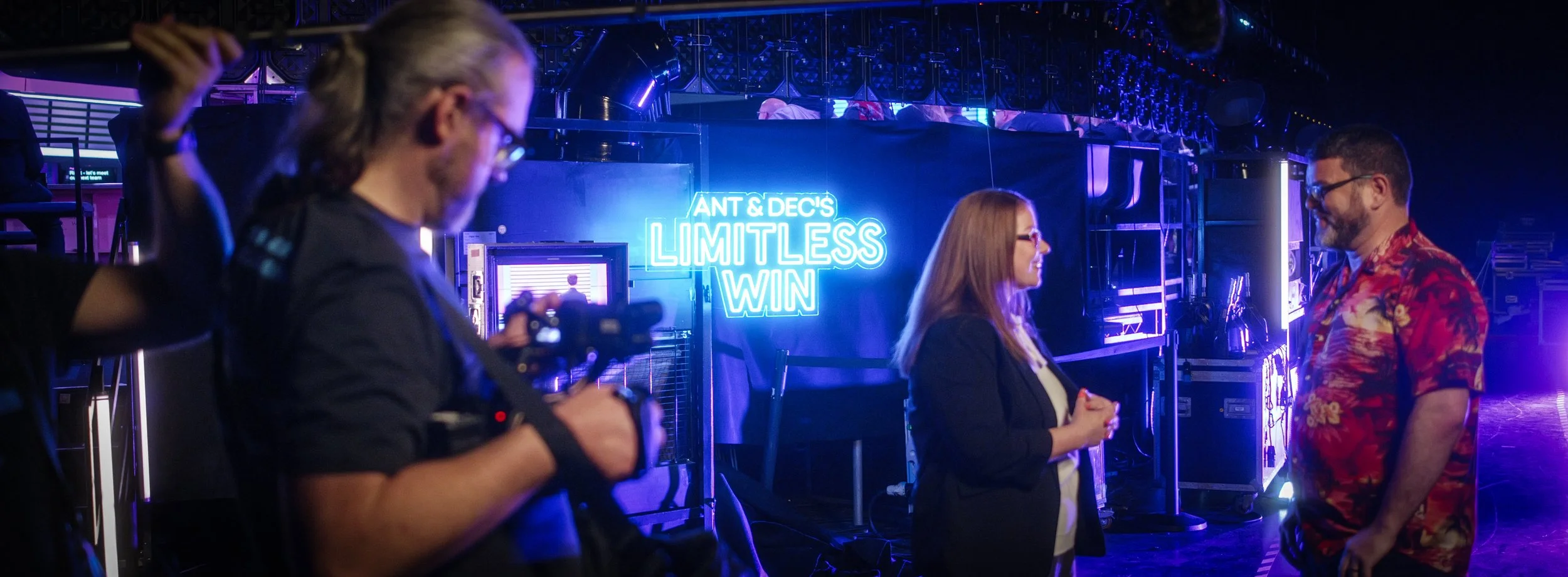 People behind the scenes at a television or stage production, with a neon sign reading 'ANT & DEC'S LIMITLESS WIN' in the background, and one person holding a camera.