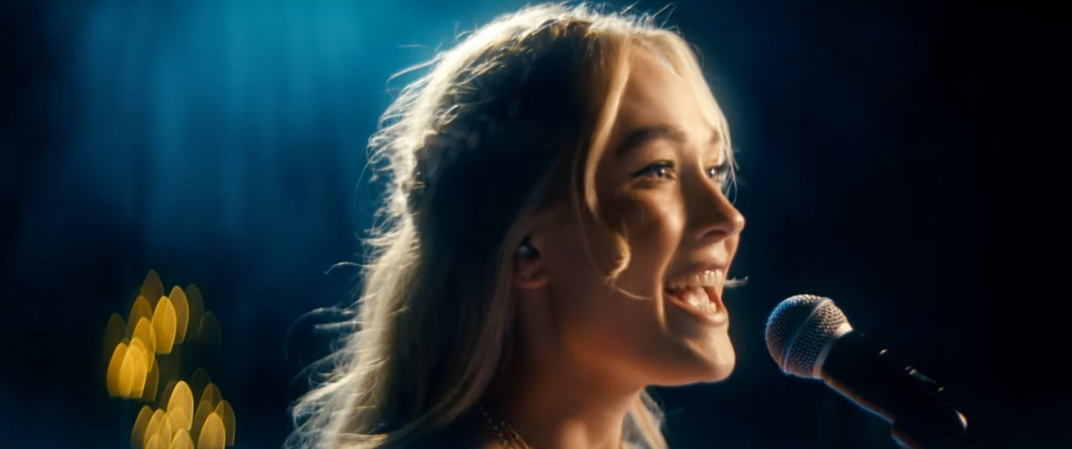 A young woman with blonde hair singing into a microphone on stage, illuminated by blue and yellow stage lights.