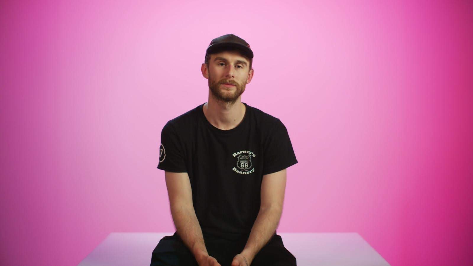 A man with light skin, a beard, and mustache sitting against a pink background. He is wearing a black t-shirt with white text and graphics, and a black cap. He is looking directly at the camera with a neutral expression.