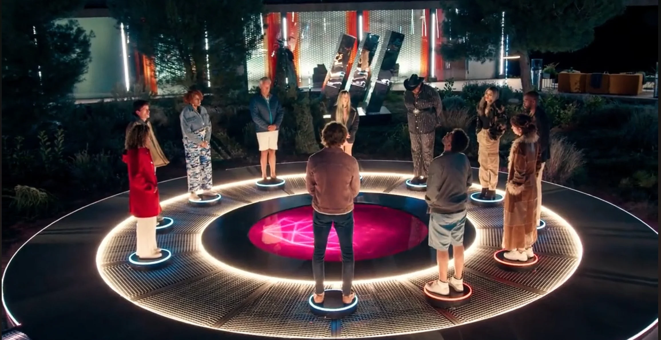 People standing on illuminated circular platforms in a semi-circle outdoors at night, with a futuristic backdrop and neon lighting.