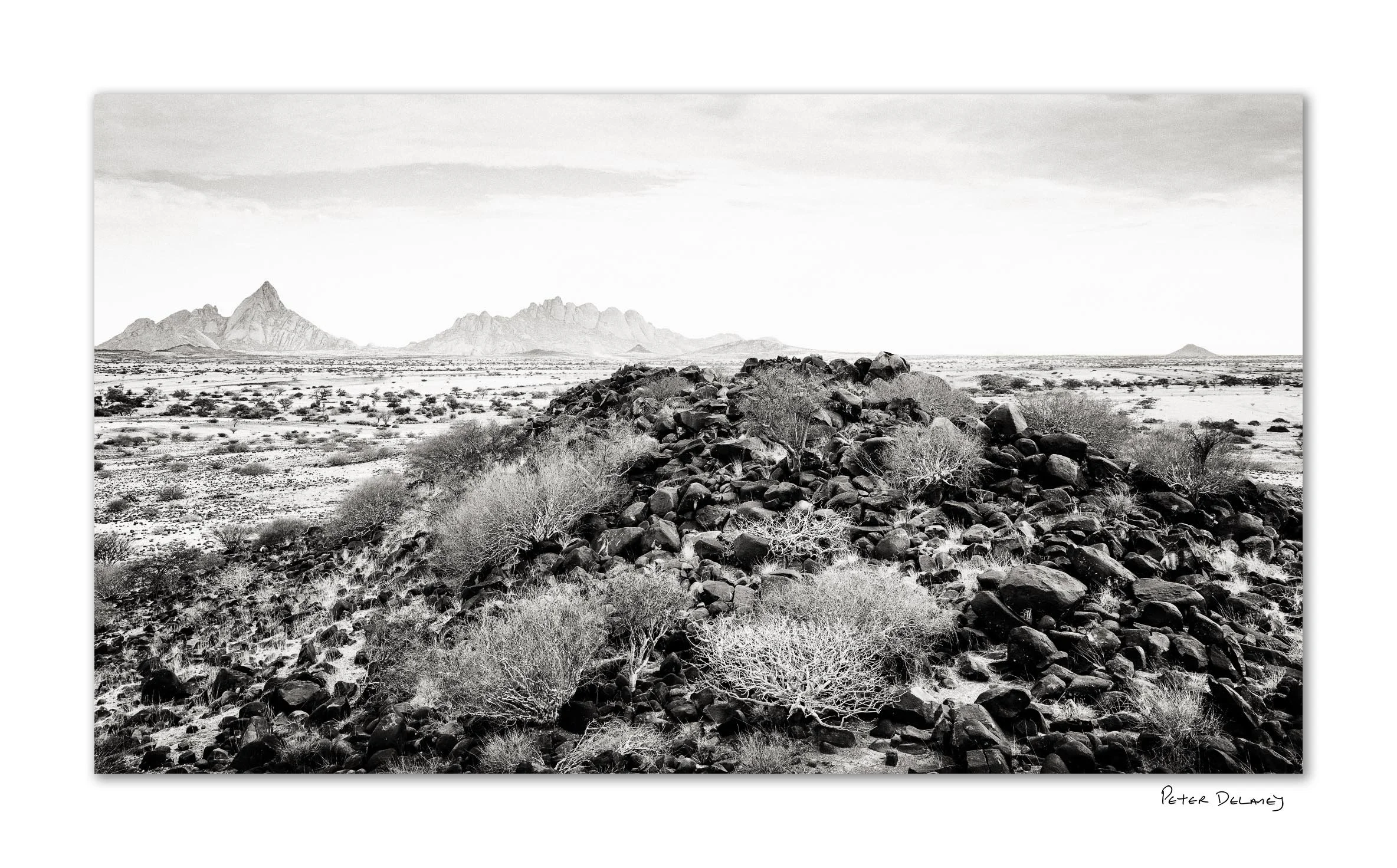 Black volcanic rocks with Spitzkoppe granite peaks in background — black and white fine art landscape print by Peter Delaney, Namibia