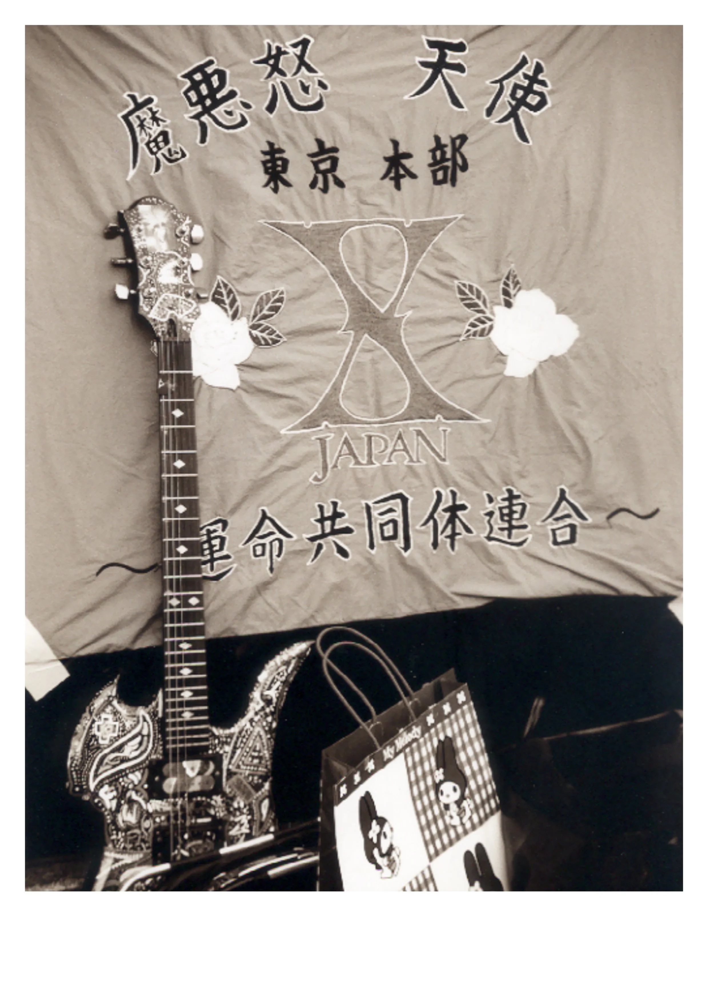 Black and white street photograph of a guitar leaning against a wall with a Hello Kitty shopping bag and a handwritten Japan sign, Yoyogi Park Tokyo 1990s, by Peter Delaney