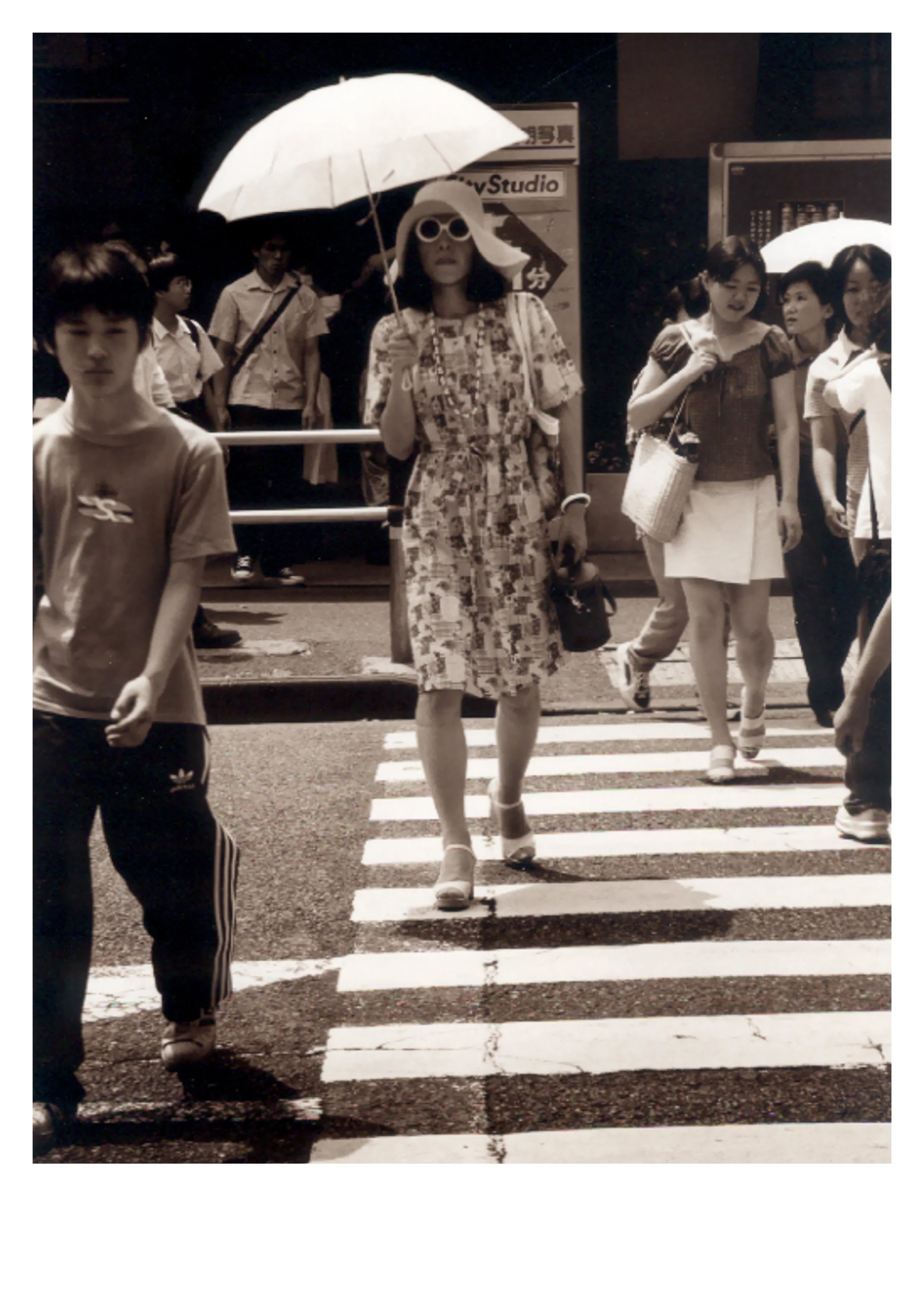 Black and white street photograph of a tall figure dressed as a woman crossing a zebra crossing in Tokyo, holding an umbrella, looking directly at the camera, by Peter Delaney