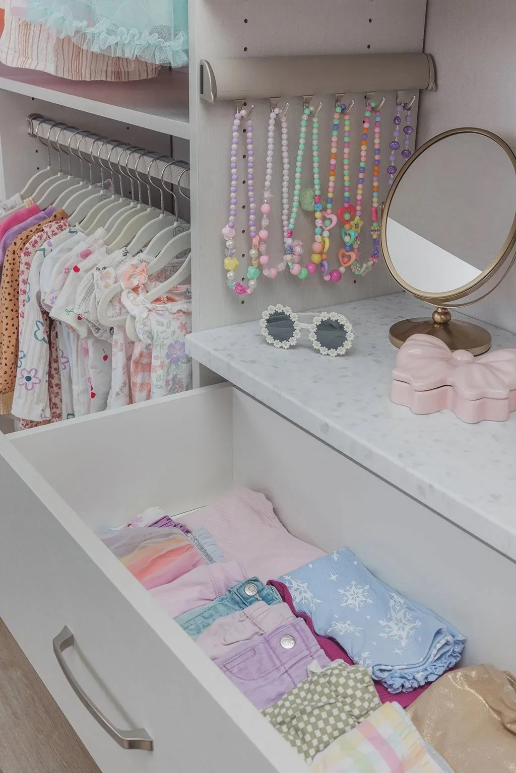  Colorful, neatly folded clothing in the open drawer of a child’s closet. The drawer has a light neutral Linen finish and brushed chrome hardware, with a white terrazzo style vanity top. Above the vanity, a slide-out jewelry organizer holds necklaces