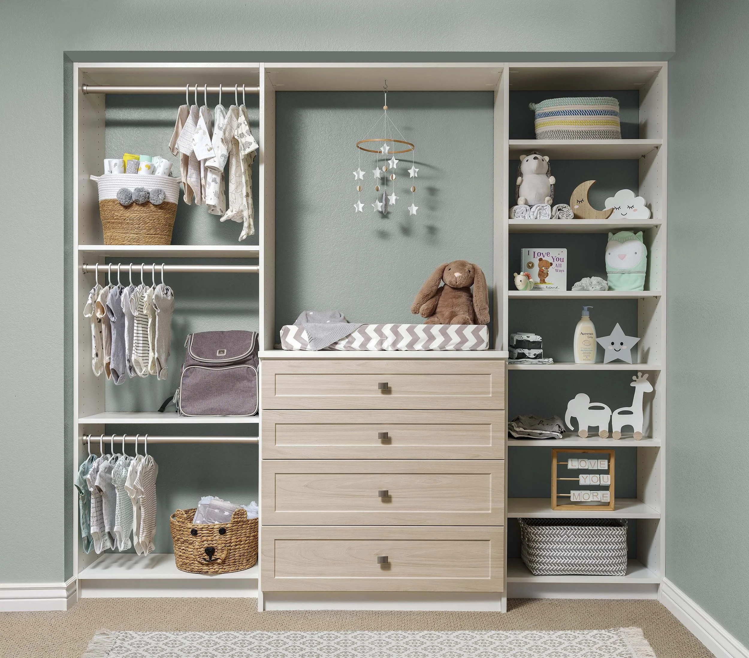 A neatly organized, soft neutral reach-in baby closet with sage green walls. White adjustable shelving and 3 levels of hanging space flank a 4-drawer dresser with white oak one-piece shaker drawer fronts and a changing table on top.