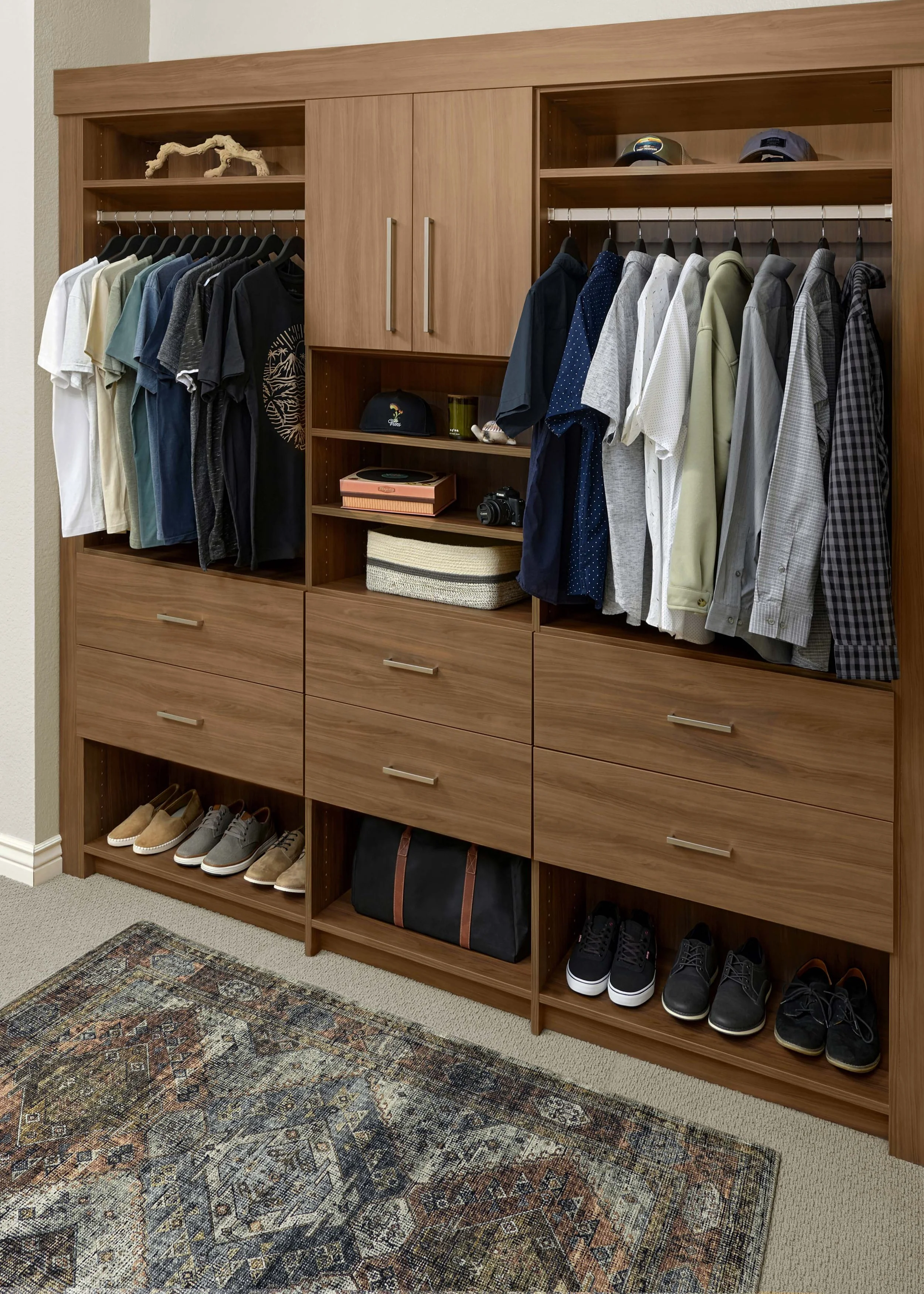 A wall of modern, built-in closet storage has a warm wood finish, Flat Panel doors, matte nickel hardware, and men's clothing and accessories neatly stored on closet rods and adjustable open shelving and in drawers and cabinets.