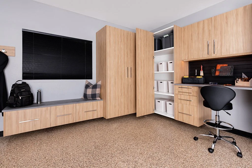 Built-in garage cabinets in a blonde wood finish are mounted along two walls of a modern garage. The design features a workbench with black slatwall tool storage, vertical storage lockers, and a built-in storage bench.