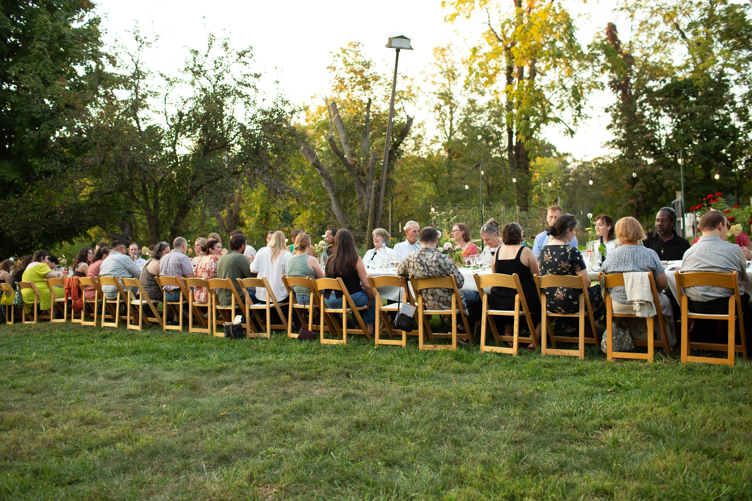 A row of 100 people sit together at a long table in a field. The sun is shining, people are smiling, lights shimmer. The atmosphere is festive and fun.