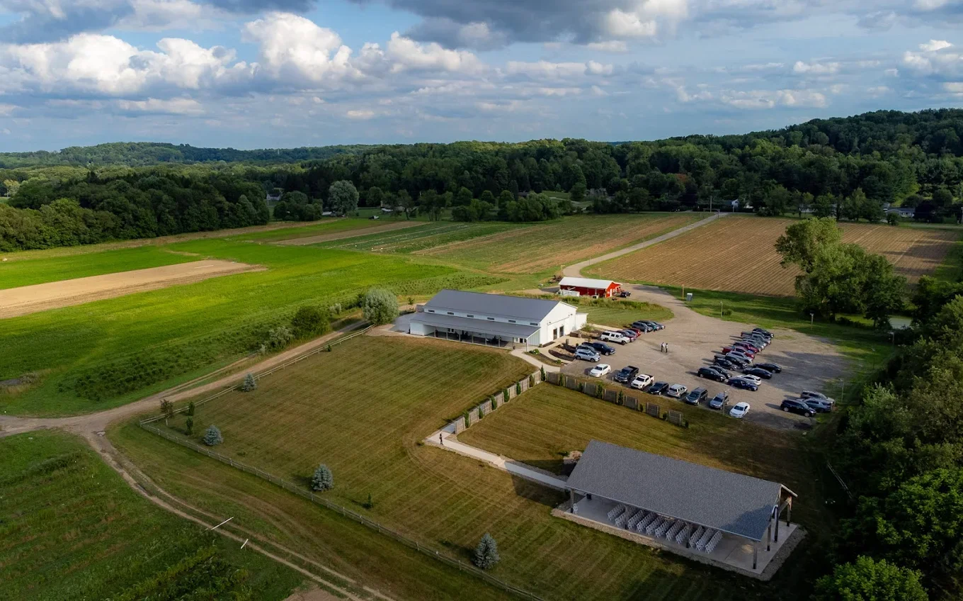 A flyover view of the Barn at Walnut Drive. The outdoor chapel sits in the shade at the bottom right in the foreground, moving to a wide view of the Barn at center with the parking lot, with green plowed fields behind and trees in the background.