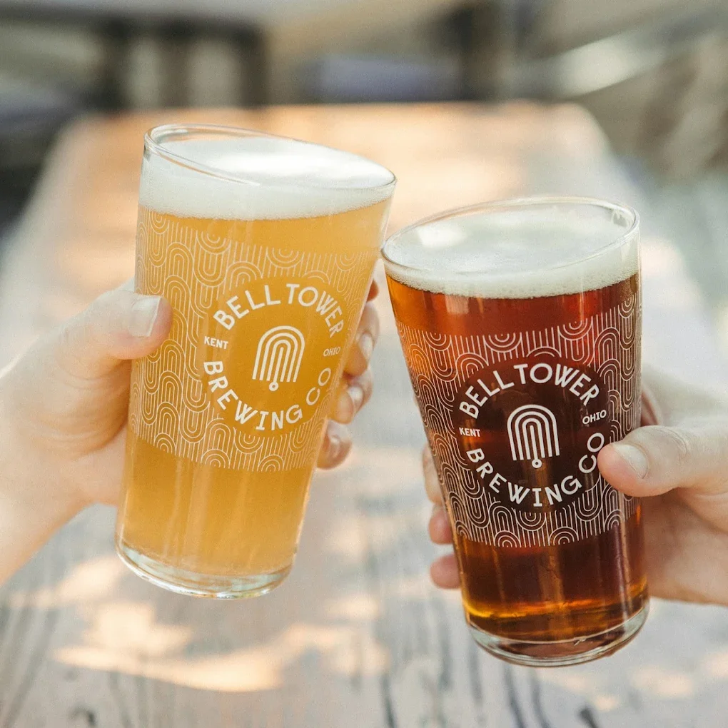 two glasses with belltower logos toast with two different shades of beer in them.