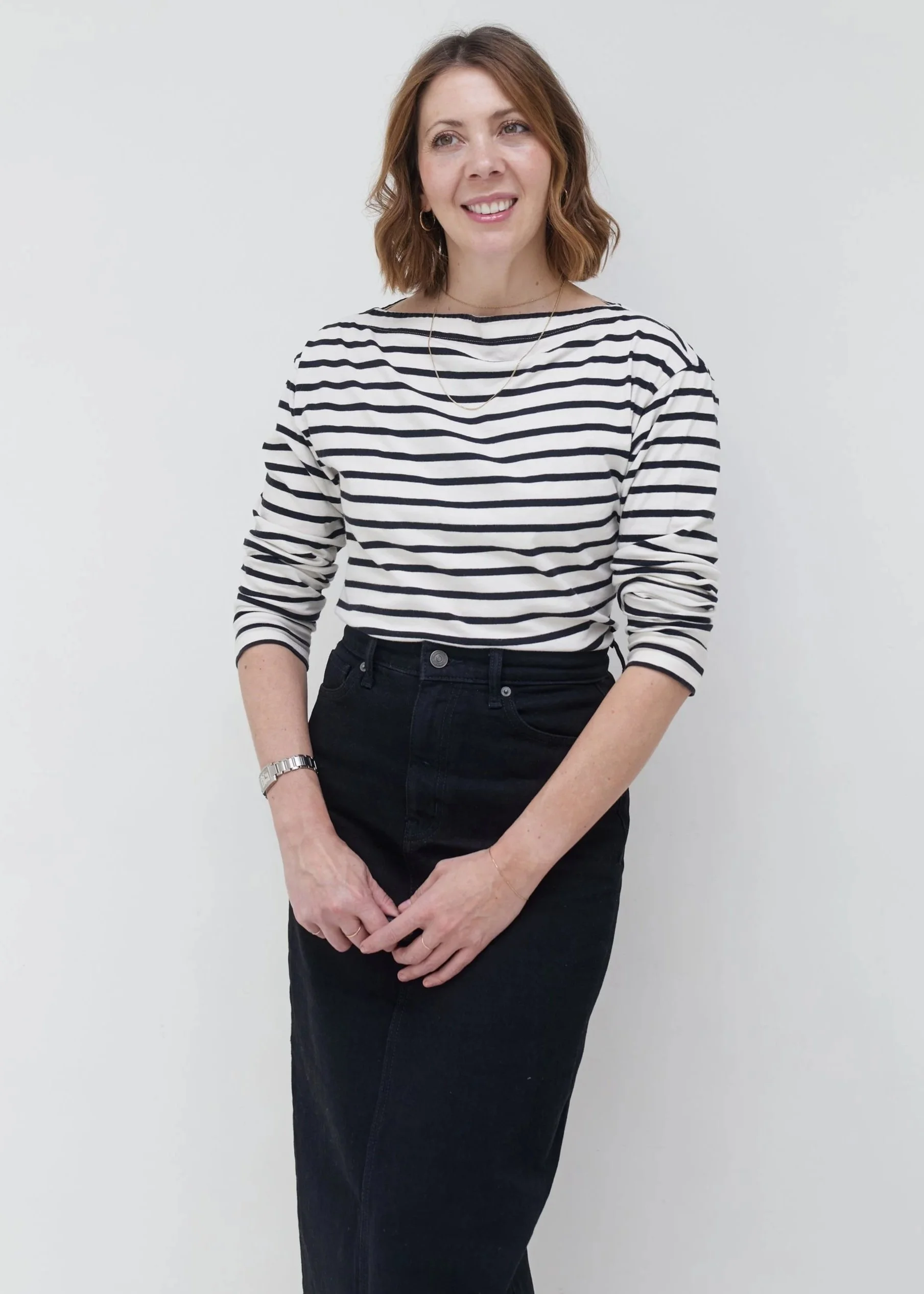 A woman with shoulder-length wavy red hair smiling, wearing a black and white striped long-sleeve shirt and black high-waisted pants, standing against a plain white background.