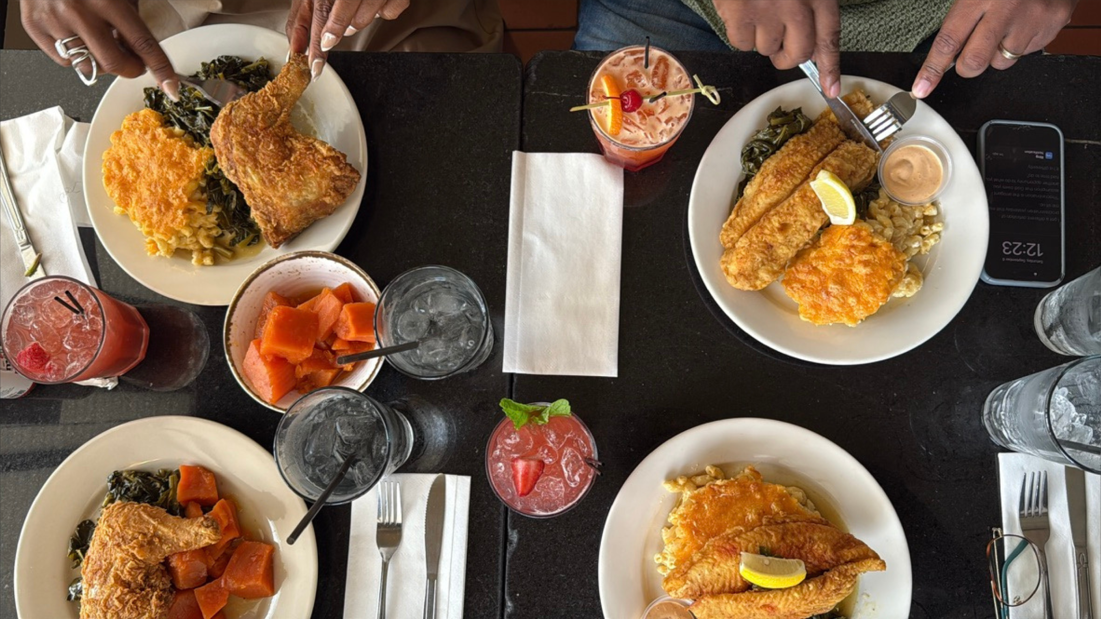 Overhead view of a soul food meal at Melba’s in Harlem with fried chicken, fried fish, macaroni and cheese, collard greens, candied yams, and colorful cocktails.