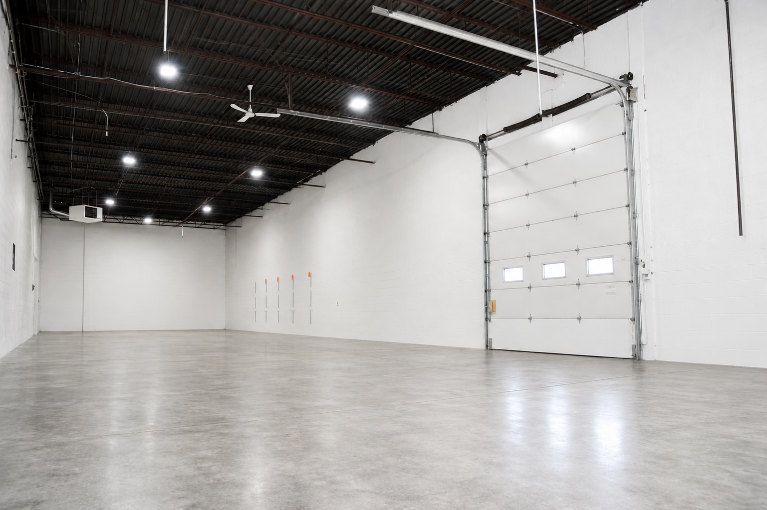 Empty warehouse with white painted brick walls, concrete floor, large garage door with small windows, ceiling fans, LED lights, and high ceiling with exposed metal roof.
