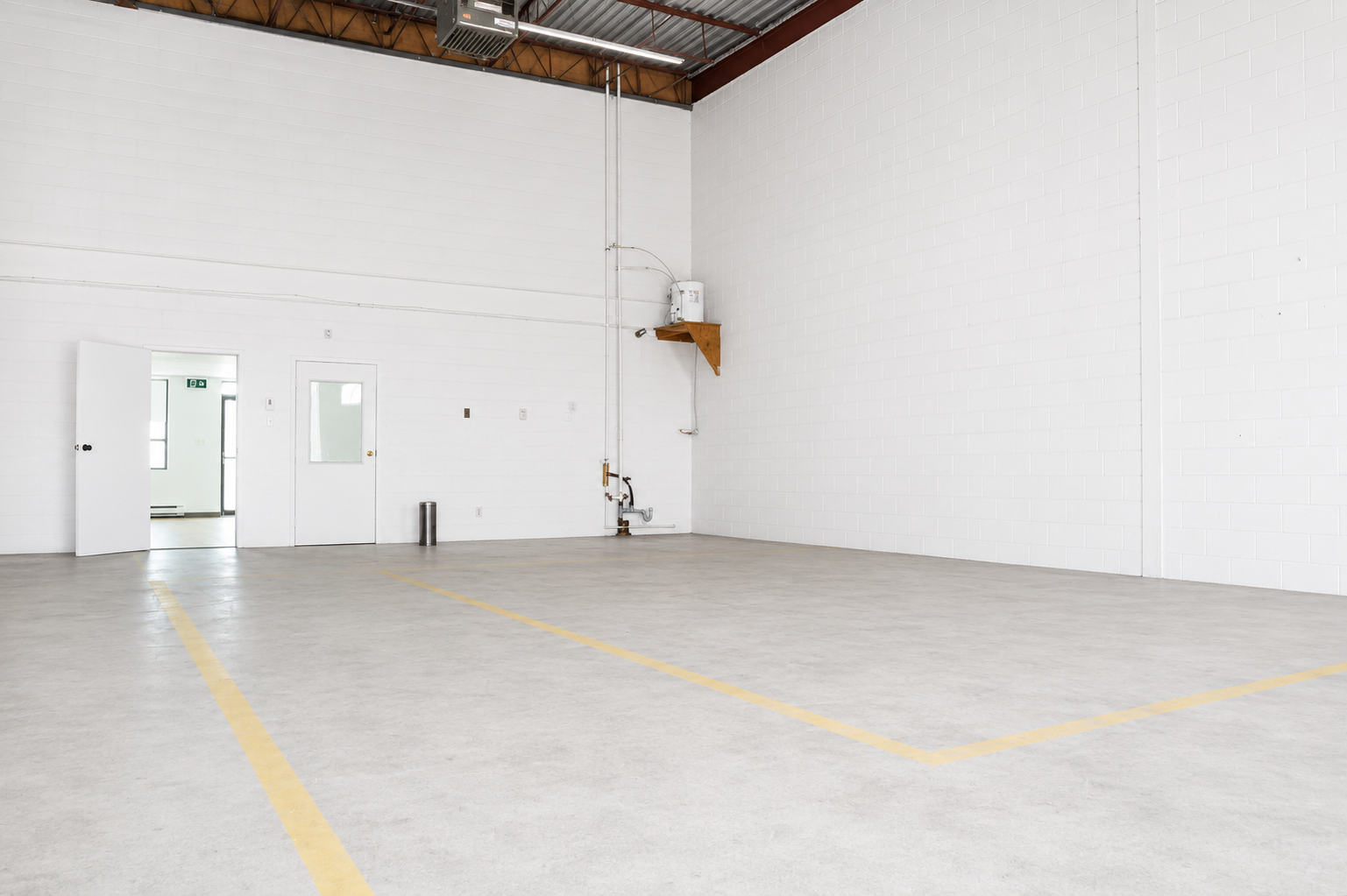 Empty indoor storage or warehouse space with white cinder block walls, concrete floor, yellow markings, and a high ceiling with metal beams and ventilation ducting.