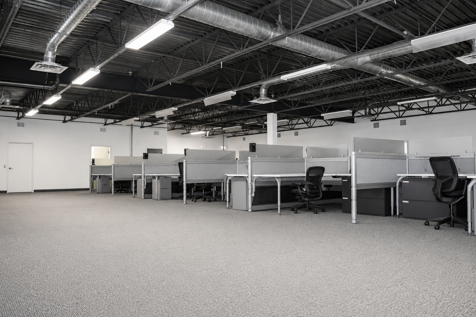 Empty office space with cubicles, office chairs, gray carpet, and industrial ceiling with exposed ducts and fluorescent lighting.