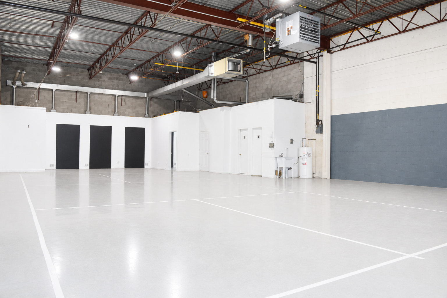 Empty indoor sports court with white lines, white and gray walls, and industrial ceiling with exposed ductwork and lighting.