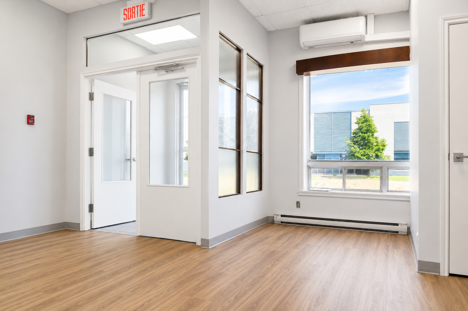 Empty room with wooden flooring, white walls, large window showing trees and buildings outside, and glass-paneled interior door.