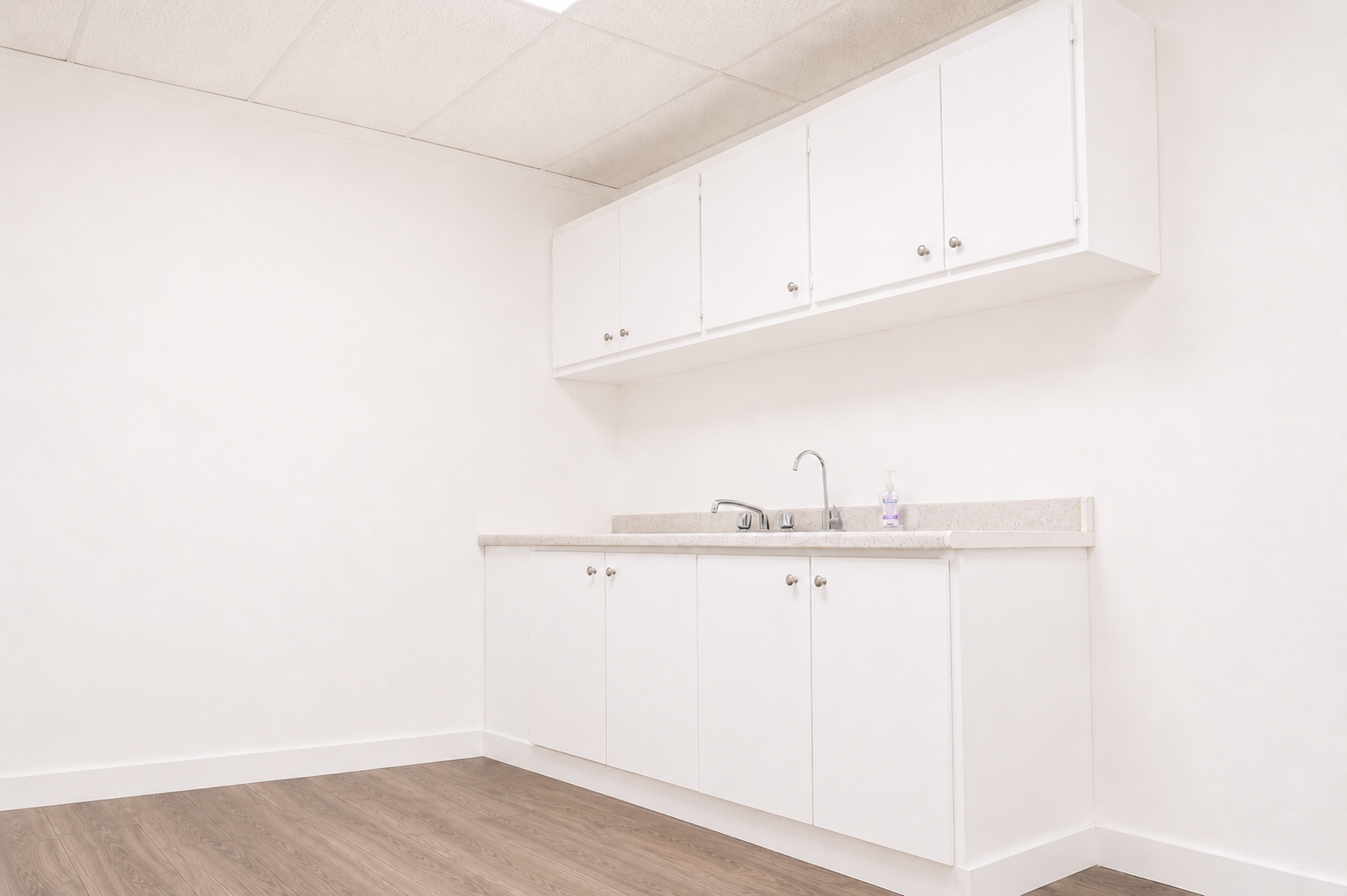 Empty white kitchenette with white cabinets, beige countertop, and a small hand soap bottle, with wood flooring and a white wall and ceiling.