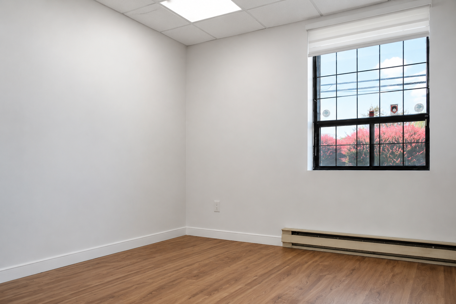 Empty room with white walls, wooden floor, a window with black frame, and a heating baseboard along the wall beneath the window.