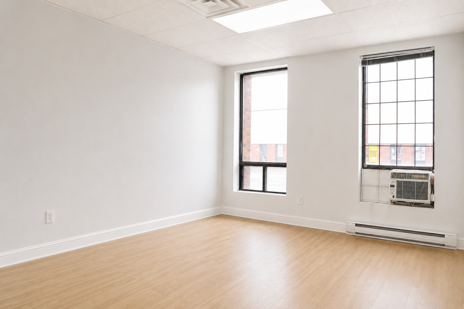 Empty room with light-colored hardwood floors, white walls, two windows with black frames, one with an air conditioning unit installed, and a ceiling with a light fixture.