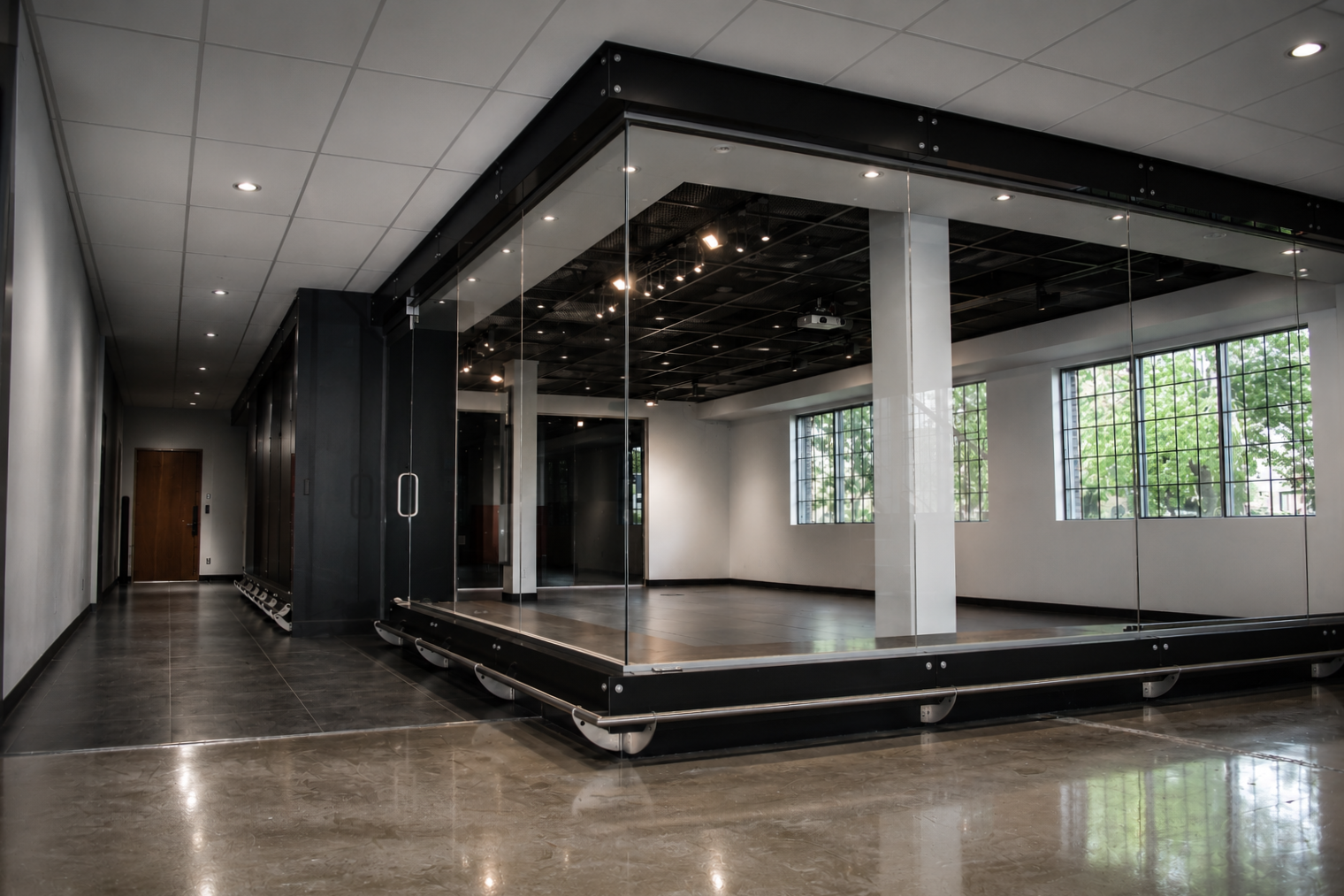 Empty dance or yoga studio with large mirrors on the wall, wooden floor, large windows letting in natural light, and black ceiling with lights.