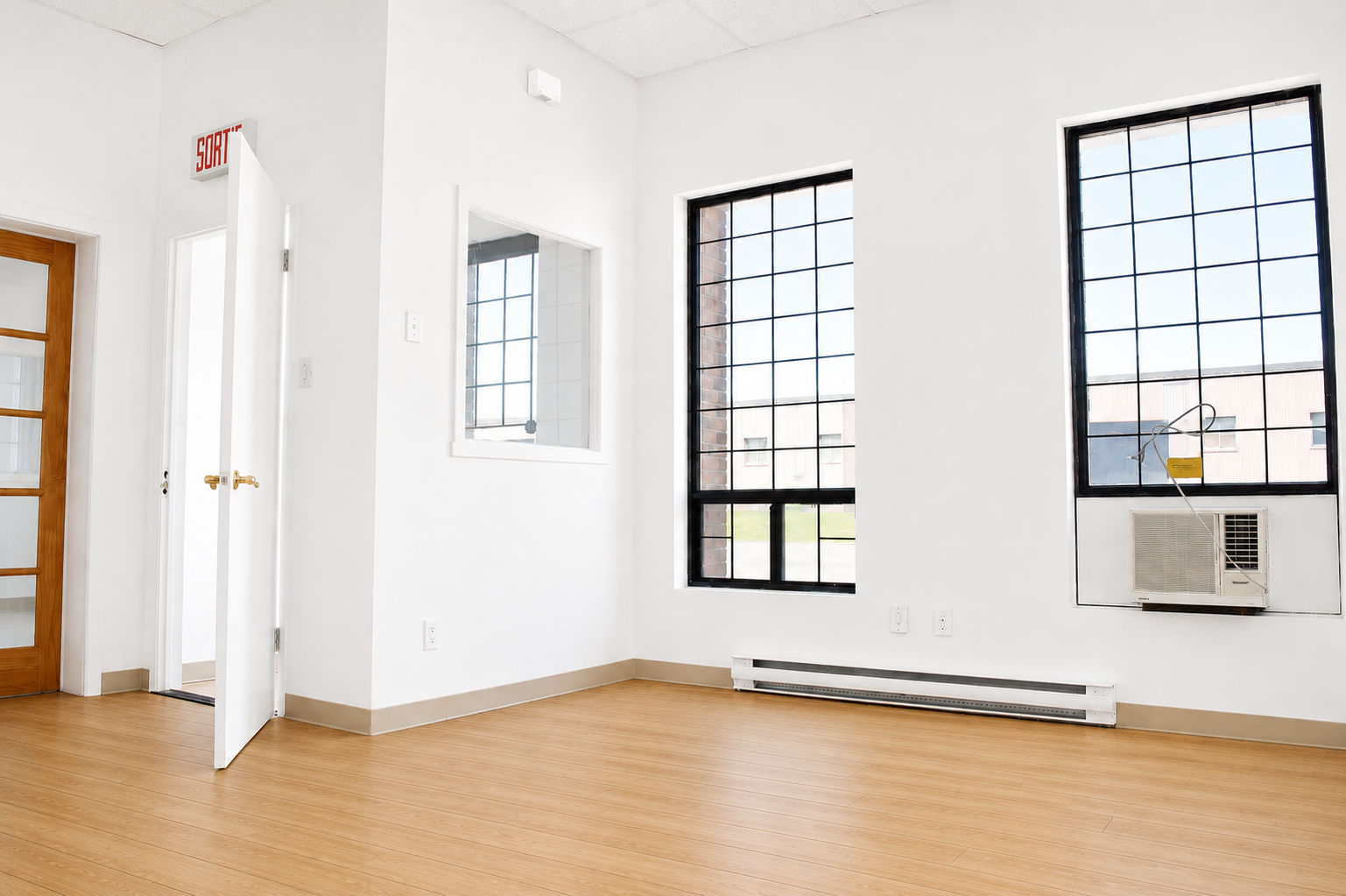 Empty room with white walls, large black-framed windows, a wood floor, and an air conditioning unit installed in one window.