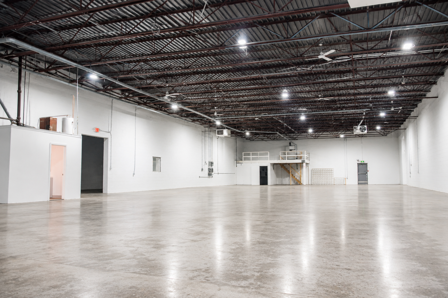 Empty industrial warehouse with high ceiling, concrete floor, and white walls, featuring small rooms and storage areas along the back wall.