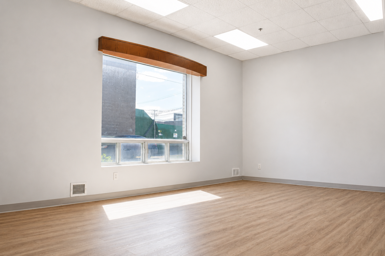Empty room with large window, light-colored wooden floor, white walls, ceiling tiles, and wooden valance above the window.