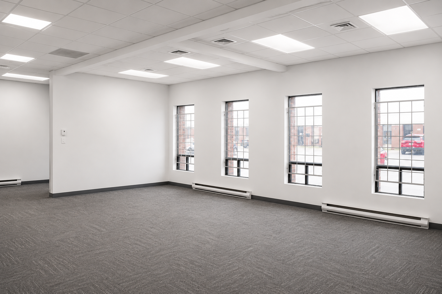 Empty office space with white walls, gray carpet, multiple windows with security bars, and ceiling lights.