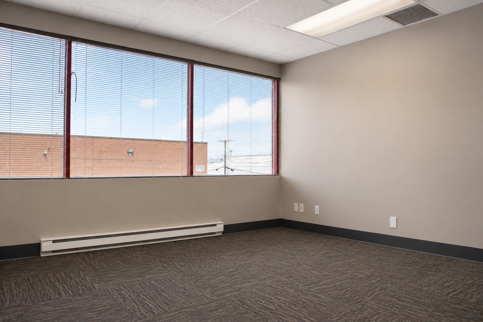 Empty office room with large window blinds, beige walls, dark carpeted floor, and electrical outlets.