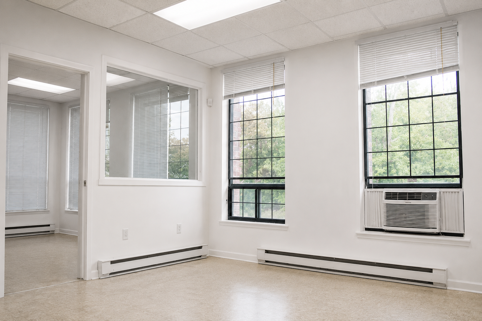 Bright office room with large windows, white walls, ceiling tiles, and window air conditioning units, overlooking trees outside.
