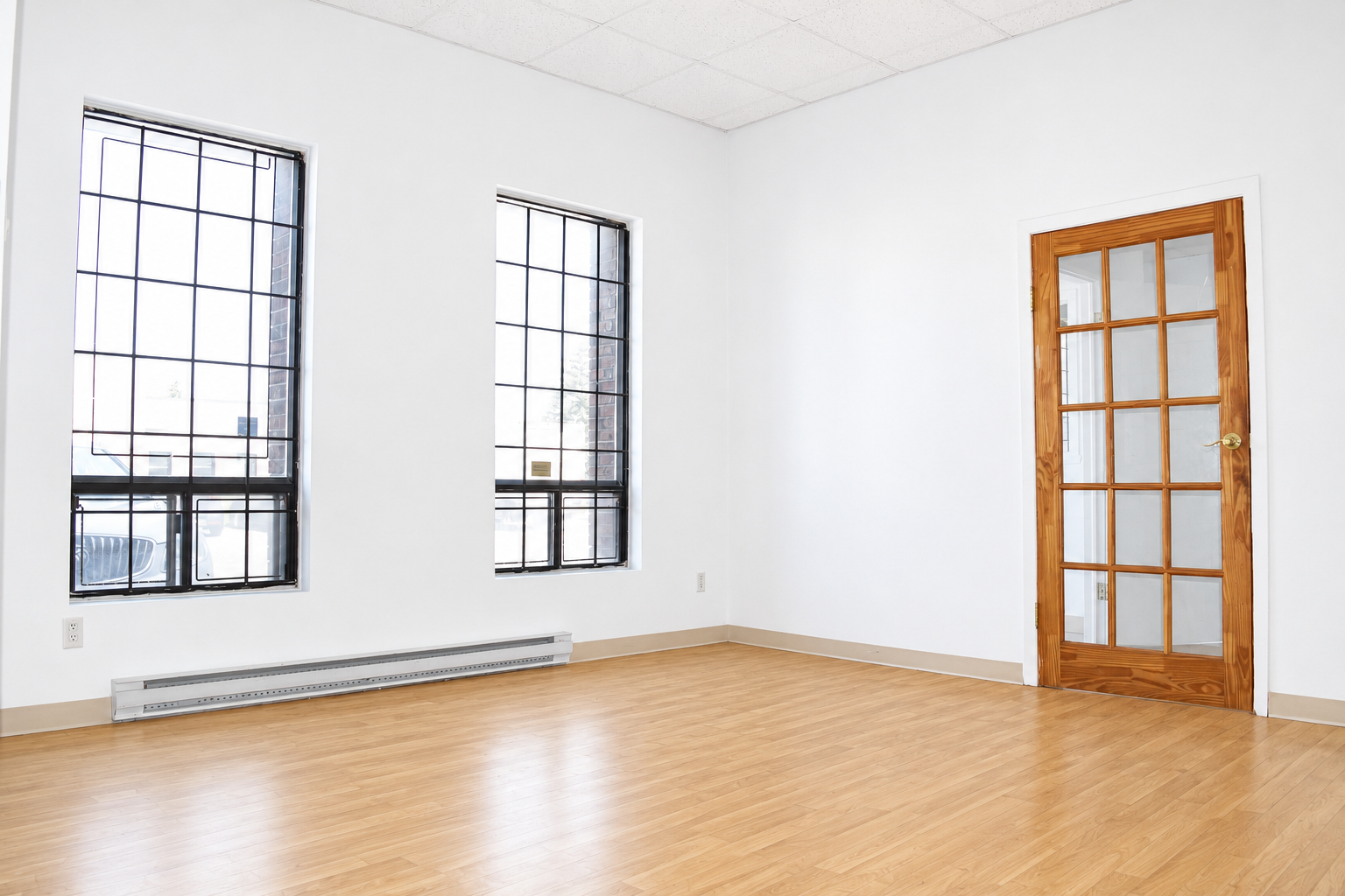 Empty room with two large windows with black frames, a wooden door with glass panels, white walls, a baseboard heater, and light wood laminate flooring.