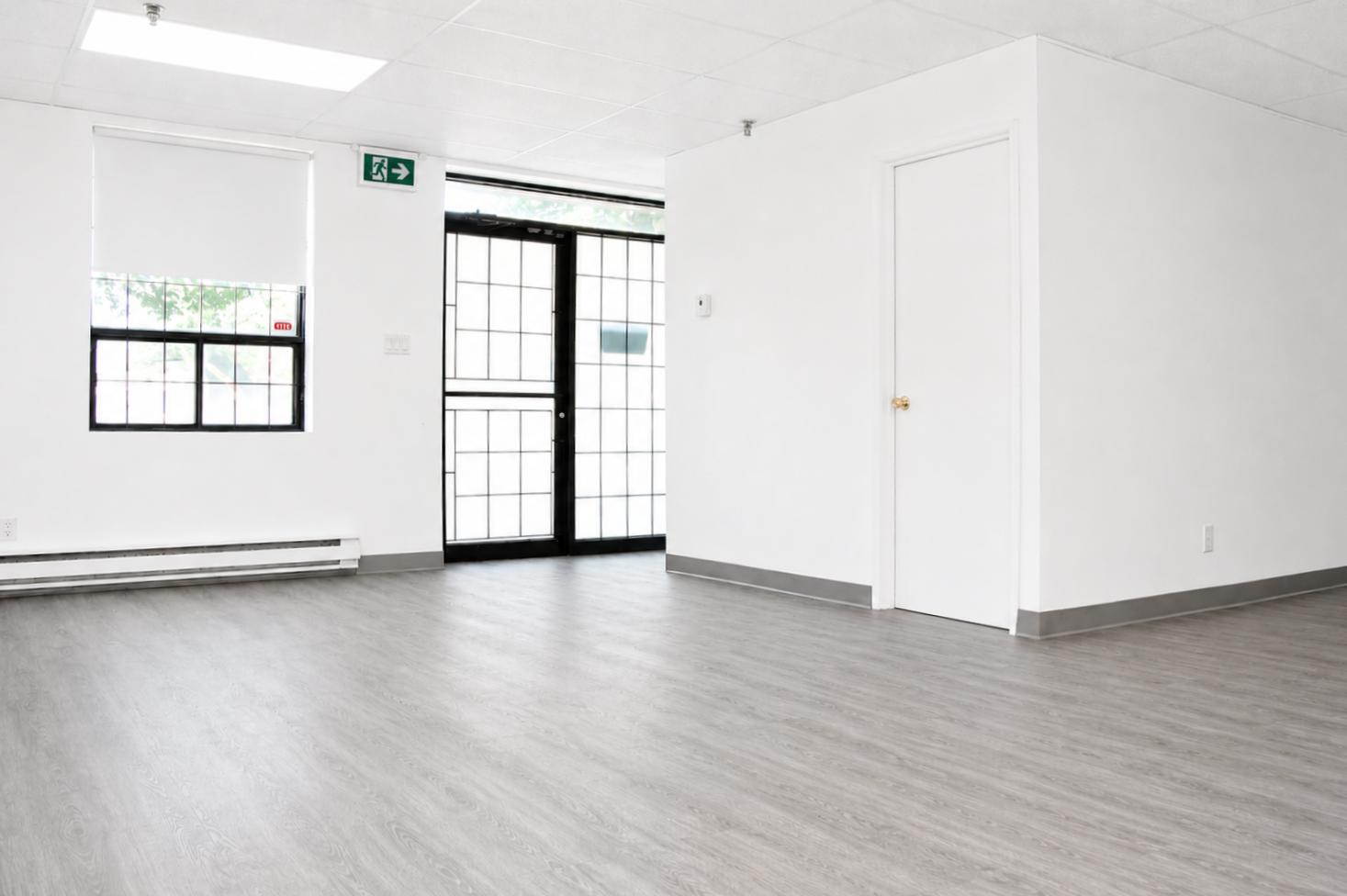 Empty room with white walls, gray floor, a window, a glass door, and an emergency exit sign.