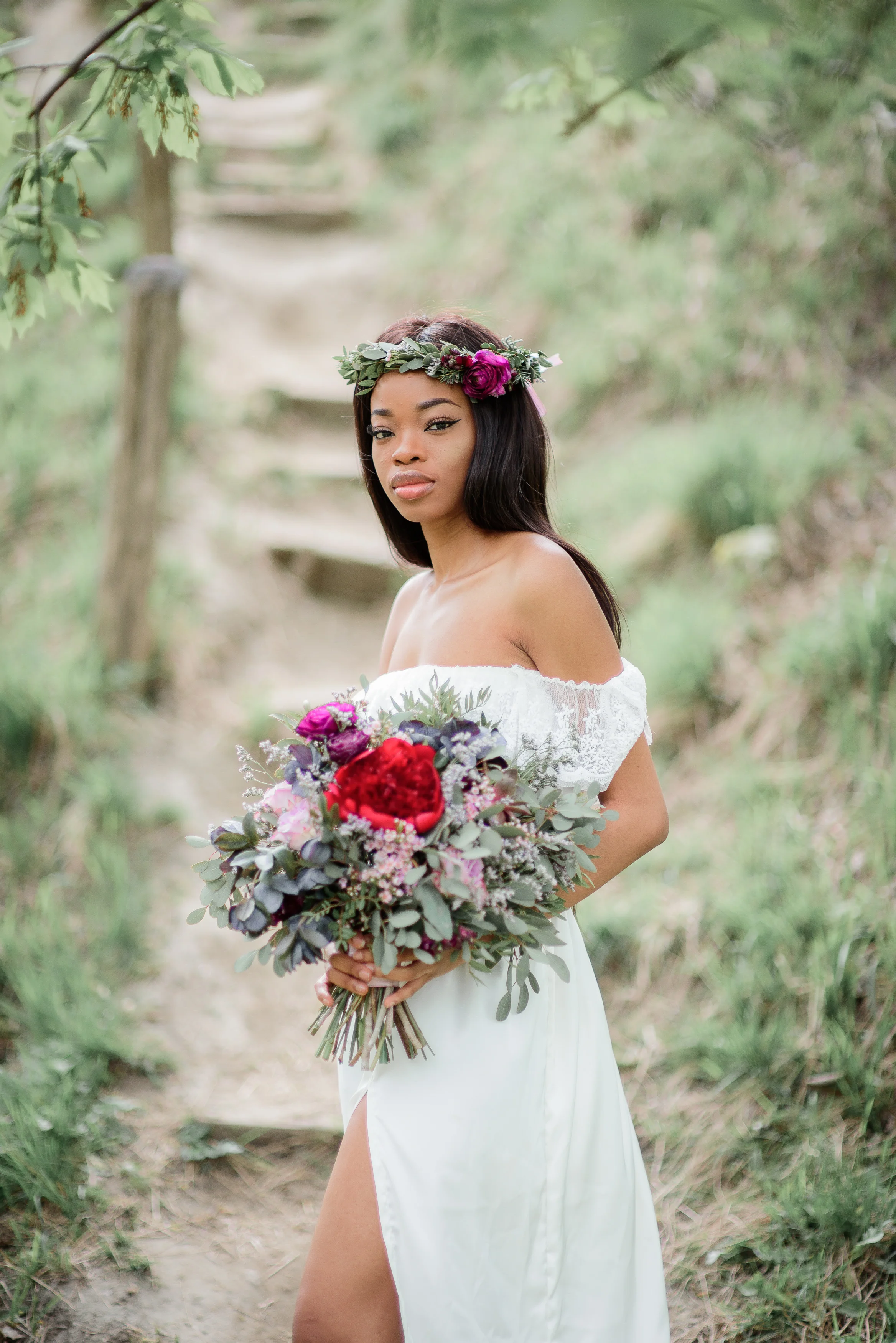 Exotic bride with dark skin in white dress with rich red bouquet stands in the park