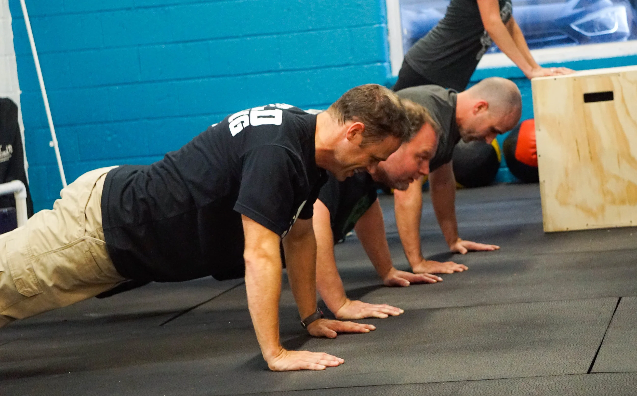 People performing push-ups on a workout mat in a gym gym with blue wall panels.