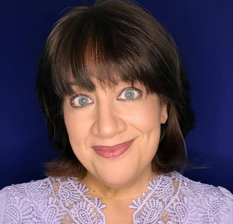 A woman with short brown hair, wearing a lavender lace top, smiling against a dark blue background.