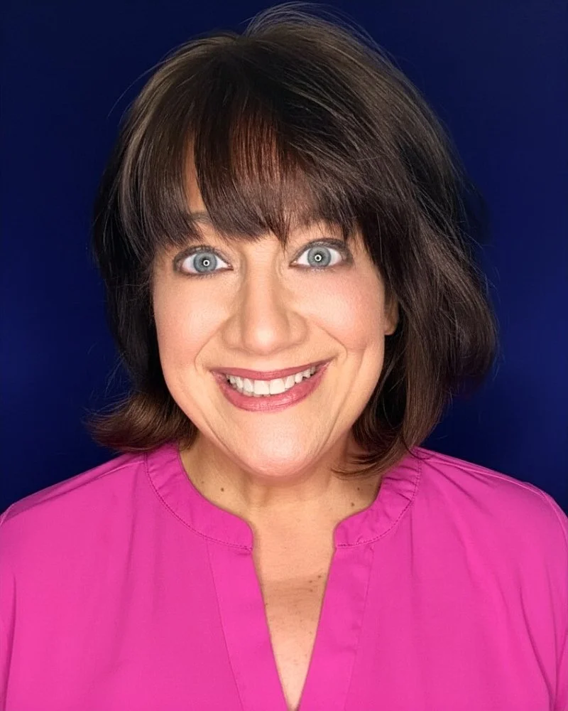 A woman with shoulder-length brown hair wearing a pink top smiling against a dark blue background.