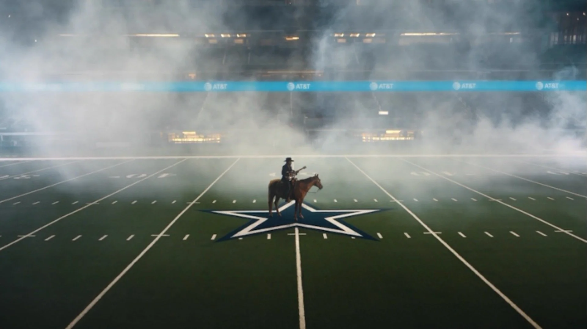 A cowboy on horseback, playing the guitar. The horse is standing on the star in the center of the field at an otherwise empty AT&T Stadium. Smoke fills the air.