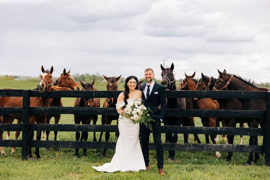  groom and bride with bouquet in front of fence with horses behind it 