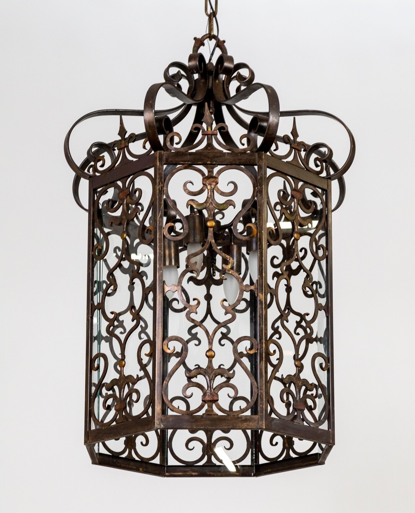 A large, hanging lantern with wrought iron scrolls, c-curves, and fleur de lis in 8 panels with clear glass. Lovely details in the canopy shape, scrolling crown, cast top loop, and subtle gold and crimson detailing. It has four, downward facing, medi