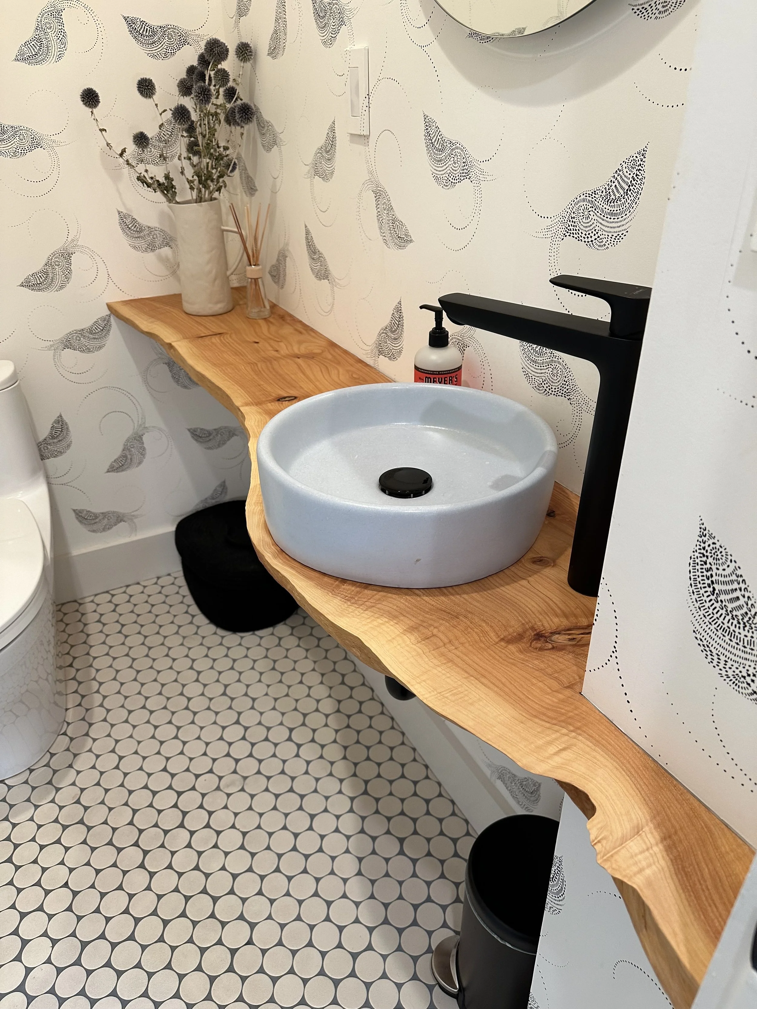 A modern bathroom with a rustic wooden countertop, a round light blue sink, black faucet, a decorative plant in a white vase, a soap dispenser, and black wastebaskets. The walls are decorated with abstract black and white patterns, and the floor has