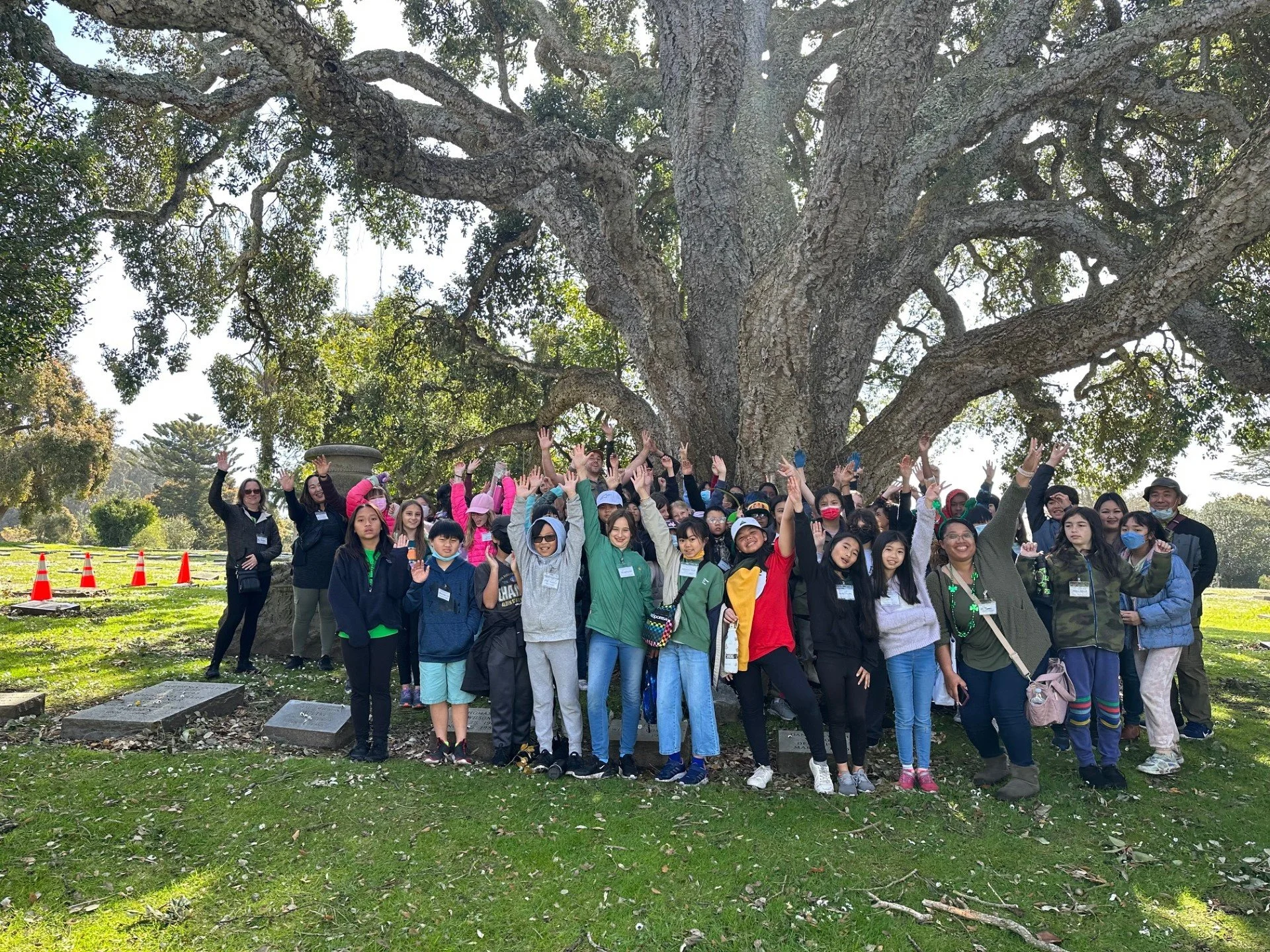 Group of children and adults standing under a large, sprawling tree in a park, posing for a photo with some raising their hands.
